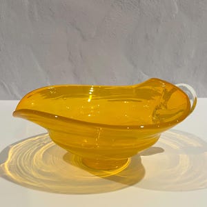 May include: A small, yellow glass bowl with a wavy rim and a clear glass handle. The bowl has a swirl design and is perfect for serving small snacks or desserts.