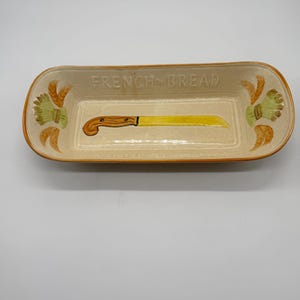 May include: Rectangular ceramic bread dish with the words "FRENCH BREAD" in the center. The dish is beige with a brown rim and features a yellow bread knife illustration. Decorative wheat and floral designs are on each end.