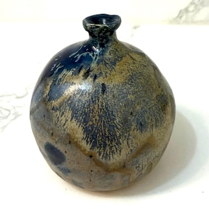Könnte beinhalten: Kleine, runde Keramikvase mit dunkelblauer und beiger Glasur. Die Vase hat einen schmalen Hals und einen bauchigen Körper. Die Glasur hat ein gesprenkeltes Aussehen mit blauen und beigen Streifen. Die Vase ist wahrscheinlich handgefertigt und eignet sich für kleine Blumen.