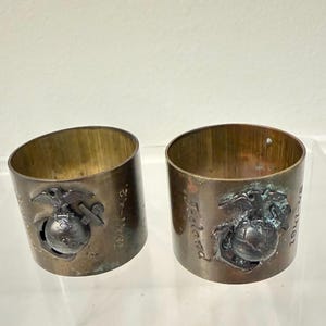 May include: Two cylindrical brass napkin rings featuring an embossed United States Marine Corps Eagle, Globe, and Anchor emblem. One ring is engraved with the text 1941-42, and the other features the name A. Toland and the text 41-42.