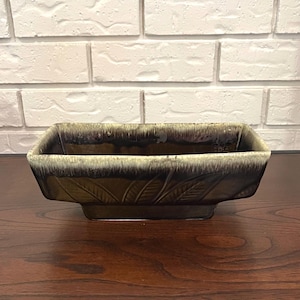 Puede incluir: Una jardinera rectangular de cerámica con un esmalte marrón oscuro y un borde marrón claro. La jardinera presenta diseños de hojas en relieve en los laterales. Se encuentra sobre una superficie de madera oscura contra una pared de ladrillos blancos.