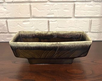 May include: A rectangular ceramic planter with a dark brown glaze and a lighter brown rim. The planter features embossed leaf designs on the sides. It sits on a dark wooden surface against a white brick wall.