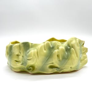 Könnte beinhalten: Eine Keramikschale mit gewelltem Rand und einem strukturierten, organischen Design. Die Schale ist hauptsächlich hellgelb-grün, mit dunkleren grünen Marmorierungen. Das Stück hat eine glänzende Oberfläche und steht auf einer weißen Oberfläche.