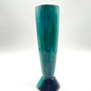 Puede incluir: Un jarrón de cerámica alto y delgado con una base acampanada. El jarrón está esmaltado en tonos de azul y verde, con una base azul más oscura.