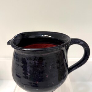 May include: A handmade, dark blue ceramic pitcher with a rounded body and a handle. The interior is a deep red color. The pitcher has a spout for easy pouring and is likely used for serving beverages.