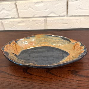 May include: Handmade ceramic plate with a dark blue center and a tan and brown rim. The plate has a unique, organic shape with raised, petal-like details on the sides. The plate is glazed with a glossy finish.