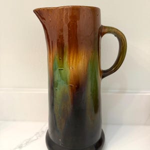 May include: A tall, ceramic pitcher with a handle and spout. The pitcher features a gradient of colors, including brown, green, and black. The pitcher has a rounded base and a smooth, glossy finish. The pitcher is likely used for serving beverages.