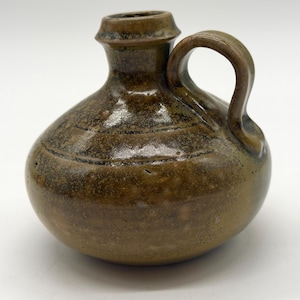 May include: A small, handcrafted brown ceramic jug with a handle. The jug has a rounded body and a narrow neck with a flared rim. Horizontal lines encircle the body. The jug is glazed with a glossy finish.
