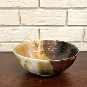 Puede incluir: Un cuenco de cerámica con un diseño único y multicolor. El cuenco presenta una mezcla de colores, incluyendo marrón, dorado, morado y blanco. Las superficies interior y exterior tienen un acabado brillante.