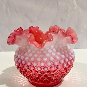 May include: A small, decorative vase in shades of pink and white. The vase features a textured, hobnail design on the body, transitioning from a deep pink base to a lighter pink and white towards the top. The rim has a ruffled edge.
