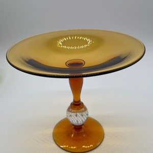 Op de afbeelding: Amberkleurige glazen voetbakschaal met een ronde basis, een decoratieve wit en goudkleurige bal en een uitlopende bovenkant. De schaal heeft een donkere rand en een kleine, cirkelvormige inkeping in het midden.