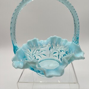 May include: A light blue glass basket with a handle. The basket has a ruffled edge and a floral design etched into the bowl. The handle has a textured, woven appearance. The basket is displayed on a clear acrylic stand.