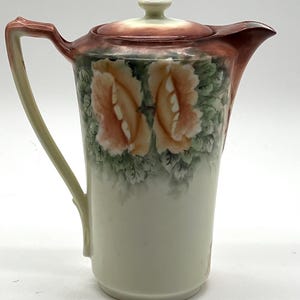 May include: A vintage porcelain pitcher with a lid, featuring a floral design of peach-colored roses and green leaves. The pitcher has a cream-colored base, a brown rim, and a curved handle.