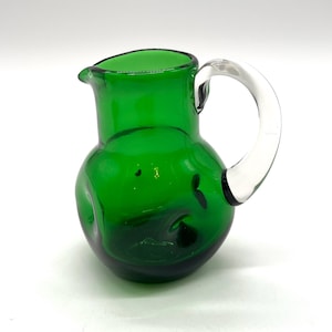 May include: A small, emerald green glass pitcher with a clear glass handle. The pitcher has a rounded body and a spout for pouring. The handle is arched and attached to the side of the pitcher. The pitcher is likely used for serving beverages.
