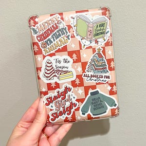 May include: A clear phone case with a pink and red checkered pattern and Christmas-themed stickers. The stickers include phrases like "Merry Christmas Ya Filthy Animal" and "Dear Santa, Send Books."