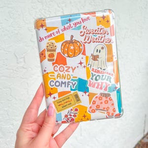 Könnte beinhalten: Eine transparente Tablet-Hülle mit einem farbenfrohen Herbst-Design. Die Hülle zeigt Grafiken von Kürbis, Kaffeetasse, Geist und Pullover sowie Text mit den Wörtern "Sweater Weather" und "Cozy and Comfy".