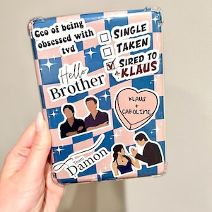 May include: A pink and blue phone case with a chequered pattern. The case has several stickers on it, including a heart with the text "Klaus + Caroline", a sticker that says "Hello Brother", and a sticker that says "Team Damon".