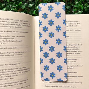 Può includere: Un segnalibro rettangolare con sfondo bianco e un motivo di fiori blu. Il segnalibro è inserito in un libro aperto. Il testo "ALBUS" è visibile in basso sul segnalibro.