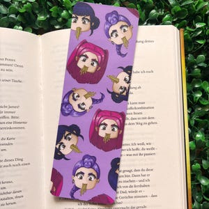 Könnte beinhalten: Ein lila Lesezeichen mit einem sich wiederholenden Muster von Cartoon-Gesichtern, jedes mit Nudeln, die aus dem Mund hängen. Das Lesezeichen befindet sich in einem offenen Buch.