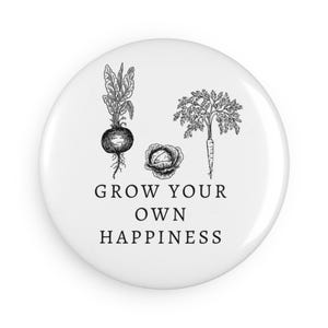 Puede incluir: Botón blanco con dibujos de línea negra de un nabo, una col y una zanahoria. El texto "Grow your own happiness" está impreso debajo de los dibujos.
