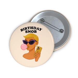 Puede incluir: Un botón redondo con el texto "BIRTHDAY SNOB" sobre una ilustración de una mujer con gafas de sol soplando una burbuja. La mujer sostiene un teléfono dorado.