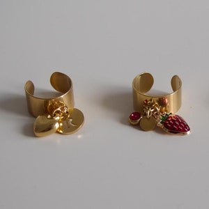 Könnte beinhalten: Drei goldene Ringe mit Anhängern. Der erste Ring hat einen Herz- und einen Buchstabenanhänger. Der zweite Ring hat eine rote Erdbeere, einen kleinen goldenen Kreis und einen roten Edelsteinanhänger. Der dritte Ring hat einen lila Kreis mit einem Halbmondanhänger.