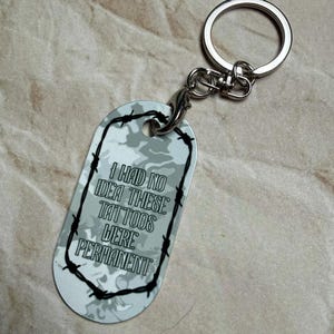May include: A silver-colored keychain with a dog tag-shaped pendant. The pendant has a camouflage pattern and the text "I HAD NO IDEA THESE TATTOOS WERE PERMANENT" in black. A black barbed wire design frames the text.