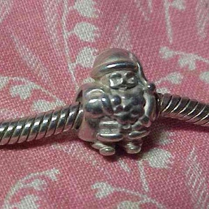May include: A silver charm featuring Santa Claus, with a hat and beard, strung on a silver bracelet. The charm is detailed with facial features and clothing. The background is a pink fabric.