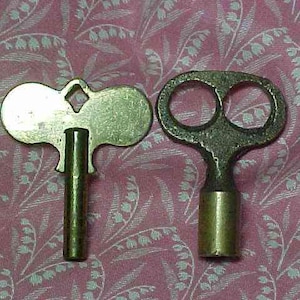 May include: Two antique brass clock keys. One key has a butterfly-shaped head with a diamond-shaped hole, the other has a double-circle head. Both keys have cylindrical stems. The keys are on a pink floral patterned background.