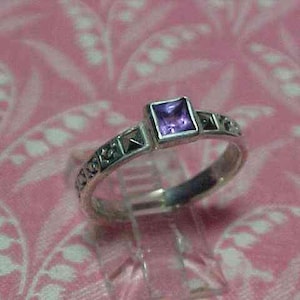 May include: A silver ring featuring a square-cut purple gemstone set in the center. The band is decorated with a series of small, raised pyramid-shaped metal accents along the sides, providing a textured appearance to the metallic surface.
