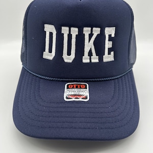 May include: A navy blue trucker hat with a white mesh back. The front of the hat has the word "DUKE" embroidered in white. The hat has a black and red label that says "OTTO WORLD FAMOUS FOAM FRONT 100%PA"