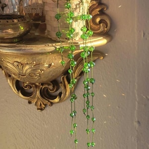May include: An ornate gold shelf displays a small woven basket with greenery and a cascade of green beads. Perfume bottles and a small gold dish are also present. The background is a neutral wall.