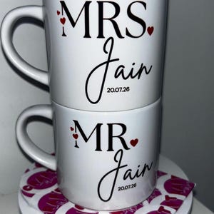 Puede incluir: Dos tazas de cerámica blanca apiladas verticalmente. La taza superior presenta el texto negro MRS. Jain con pequeños corazones rojos. La taza inferior presenta el texto negro MR. Jain con pequeños corazones rojos. Ambas incluyen la fecha 20.07.26.