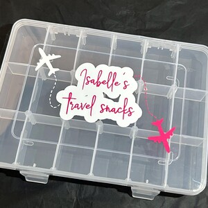 Puede incluir: Un organizador de snacks de viaje de plástico transparente con múltiples compartimentos. La tapa presenta una pegatina blanca con el texto "Isabelle's travel snacks" y gráficos de aviones en blanco y rosa. El organizador está diseñado para guardar snacks.