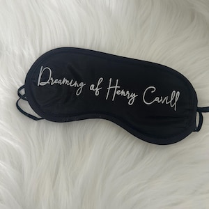 以下が含まれることがあります： 「Dreaming of Henry Cavill」の白文字が入った黒いスリープマスク。マスクは曲線的で、黒いストラップが付いています。背景はふわふわとした白い表面です。