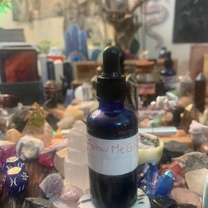 May include: A dark blue glass bottle with a black dropper, labeled "Follow Me Girl" in pink. The bottle sits on a white and gray stone, surrounded by crystals and other decorative items. The bottle contains a dark liquid.