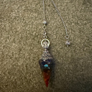 May include: A silver pendulum with a cone-shaped crystal pendant. The pendant has layers of colourful stones, including red, orange, blue, and clear. The pendulum is suspended by a delicate chain with decorative beads.