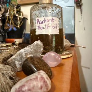 Puede incluir: Una botella de vidrio vintage con tap&oacute;n de corcho, etiquetada como "Radiant Health oil". La botella se encuentra sobre una superficie de madera con varias piedras pulidas en tonos rosa, marr&oacute;n y transparente. En el fondo se ve una colecci&oacute;n de joyas y otros objetos.