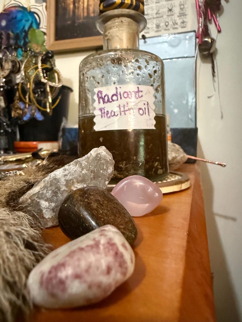 Puede incluir: Una botella de vidrio vintage con tap&oacute;n de corcho, etiquetada como "Radiant Health oil". La botella se encuentra sobre una superficie de madera con varias piedras pulidas en tonos rosa, marr&oacute;n y transparente. En el fondo se ve una colecci&oacute;n de joyas y otros objetos.