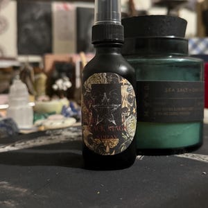 Può includere: Un flacone spray nero con un tappo trasparente e un'etichetta che recita "Black Magic Spray". Accanto, una candela verde in un barattolo di vetro con coperchio nero. Lo sfondo è sfocato.