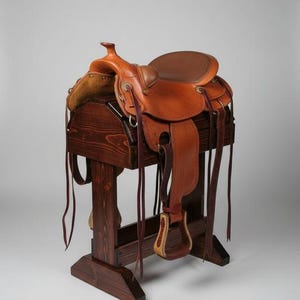 May include: A brown leather saddle with stirrups and straps rests on a dark wooden saddle stand. The saddle has a rounded seat and a horn. The stand is made of stained wood.