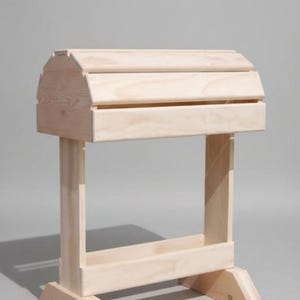 May include: A light-colored wooden saddle stand with a curved top and a rectangular base. The stand is constructed from multiple pieces of wood, with a simple, functional design. The stand is designed to hold a saddle.