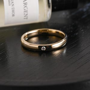 Könnte beinhalten: Ein polierter goldfarbener Bandring mit einem einzelnen kleinen, runden, klaren Edelstein, der bündig in die Mitte der Metalloberfläche eingelassen ist. Der Ring hat ein glattes, abgerundetes Profil und eine reflektierende Oberfläche.