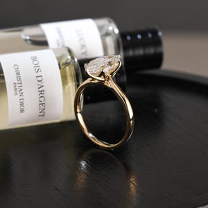 May include: A gold engagement ring featuring a pear-cut diamond. The ring is set on a slender band. Two perfume bottles are visible in the background, adding to the composition.