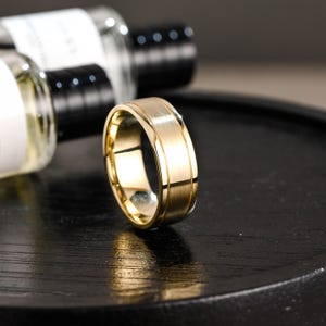 Könnte beinhalten: Ein goldfarbener Ring mit gebürsteter Oberfläche und poliertem Innenband. Der Ring hat eine flache Oberseite und Seiten mit gerilltem Design. Der Ring wird auf einer dunklen Oberfläche präsentiert.