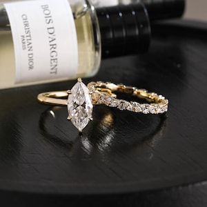 Könnte beinhalten: Ein goldener Verlobungsring mit einem großen, marquise-geschliffenen Mittelstein und ein passender goldener Ehering, der mit einer durchgehenden Reihe kleiner marquise-geschliffener Steine besetzt ist. Die Ringe liegen vor einer Flasche mit dem Etikett Bois D'Argent.