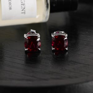 May include: A pair of stud earrings featuring deep red, cushion-cut gemstones set in polished silver-toned metal prongs. The earrings are positioned in front of a partially visible glass bottle with a label containing the text DIOR and GENT.