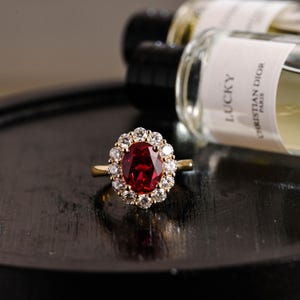 May include: A gold ring with a large, oval-cut, deep red gemstone encircled by smaller, clear gemstones. The ring is set against a dark surface, with perfume bottles visible in the background.