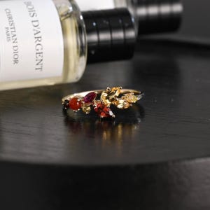 May include: A gold ring featuring a cluster of marquise and round-cut gemstones in shades of red, orange, and yellow. The stones are arranged in a leaf-like pattern along the band, showcasing deep garnet, bright citrine, and warm amber hues.