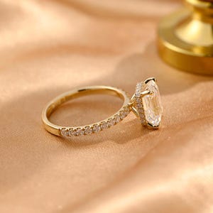 Op de afbeelding: Een gouden ring met een grote, vierkante diamant die is gezet in een halo van kleinere diamanten. De ring heeft een dunne band die ook is bezet met kleinere diamanten.